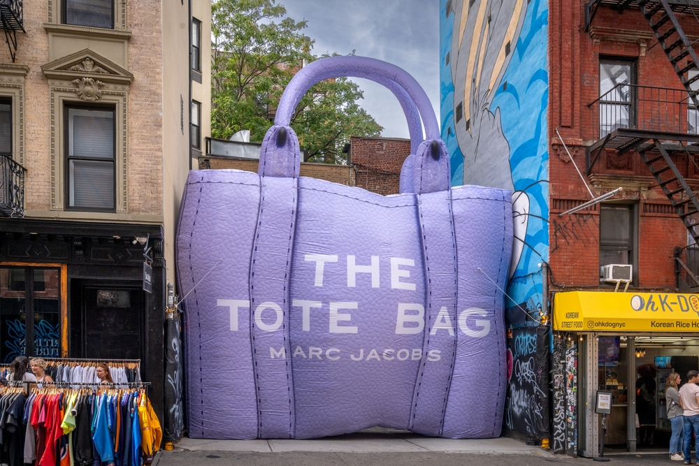 A large purple leather bag with the words 'The Tote Bag by Marc Jacobs' is squeezed between two buildings in New York.