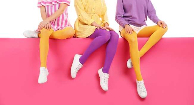 A row of three people sitting on a pink block wearing bright pink, yellow and purple outfits with contrasting yellow and purple tights and white shoes.